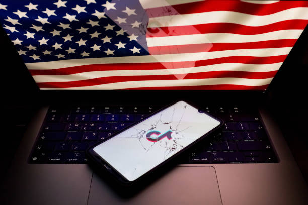 In this photo illustration, the TikTok logo is being displayed on a phone screen seen through broken glass, with an American flag appearing on a screen in the background in Athens, Greece, on April 25, 2024. (Photo by Nikolas Kokovlis/NurPhoto via Getty Images)